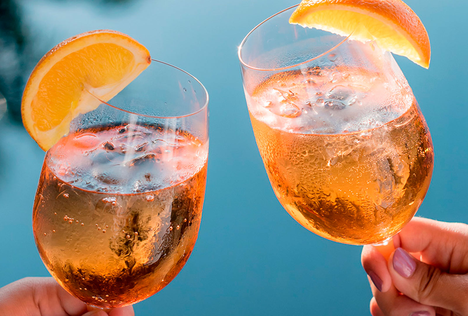 Dos manos sosteniendo vasos con líquido y rodajas de naranja